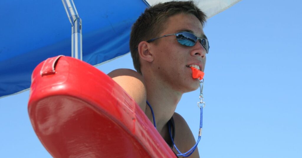 A life guard holding a life preserver with a whistle in his mouth. This is among the best summer jobs for teens.